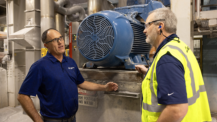 Two people stand by an electric motor, they are wearing safety gear and discussing the motor.