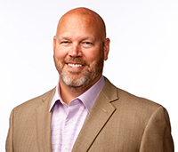 Smiling bald man with a grey bird and moustache wearing a brown suit jacket and a pink shirt.