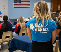 Woman stands as she listens to a presentation in a classroom, her shirt says "Human Powered. Avista" on the back in white letters.