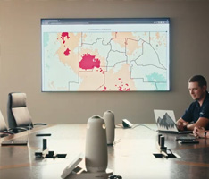 In a meeting room, a map is shown on the screen while people sit around the table.