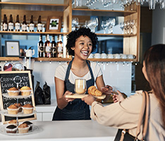 Worker at coffee shop hands a customers a tray with a pastry and a coffee, there are more fresh pastries on the counter display